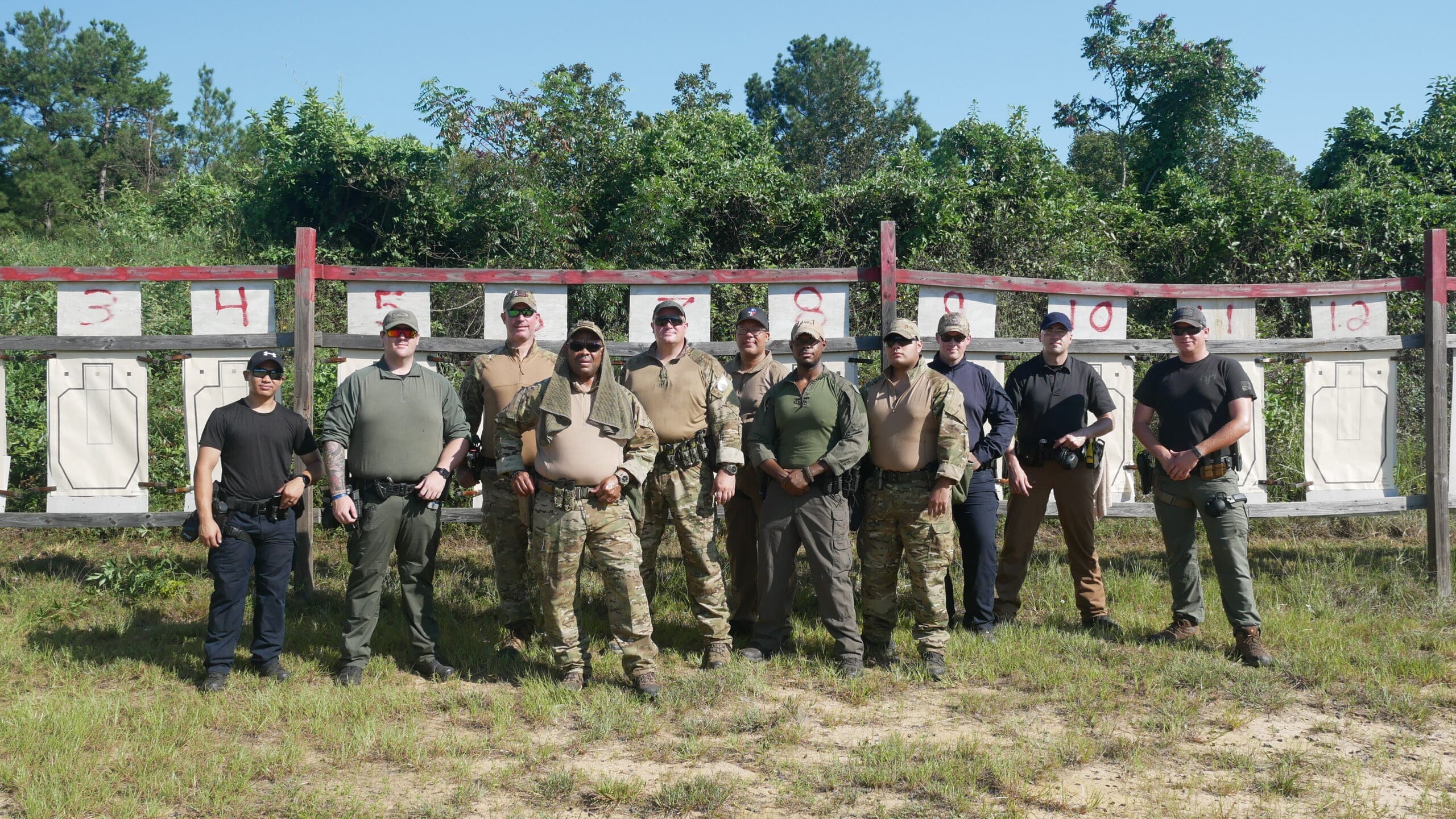 A group of twelve men in tactical and casual attire stand in a line on a grassy field.
