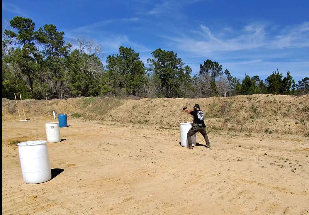 A person with a handgun practices shooting behind cover at a training range, surrounded by barrels and greenery under a clear blue sky.